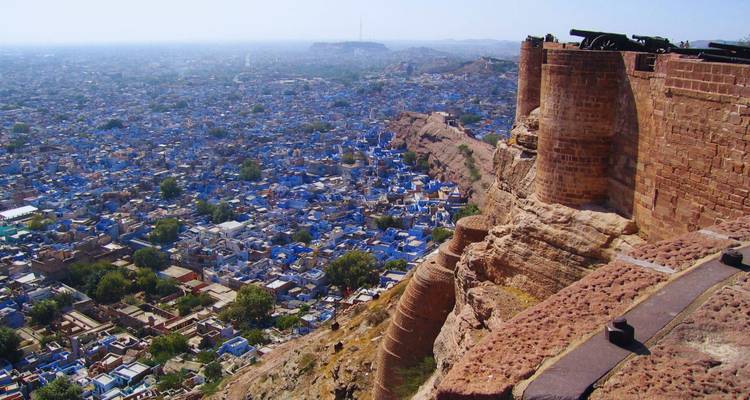 View from fort ramparts over a sprawling blue city with cannons lined along reddish stone walls.