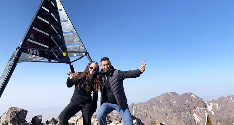 Mannelijke en vrouwelijke klimmer poseren met vredestekens naast de metalen toppiramide onder een heldere blauwe hemel.