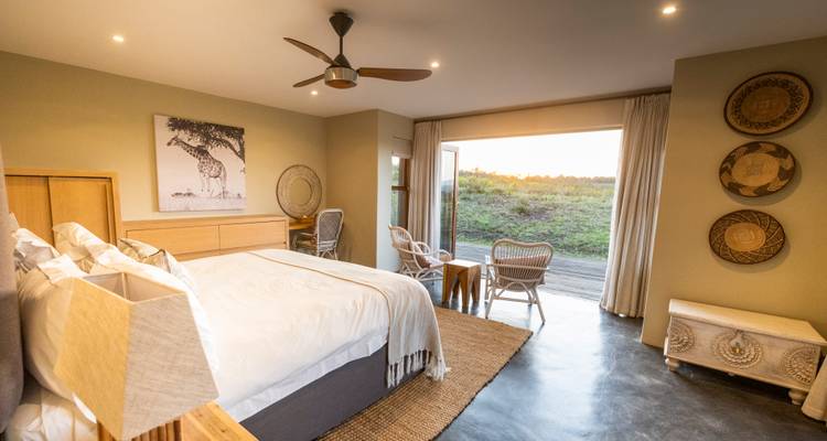 Une chambre de lodge élégante avec un grand lit, des chaises en osier et des portes ouvertes menant à une terrasse surplombant des plaines vertes au coucher du soleil.