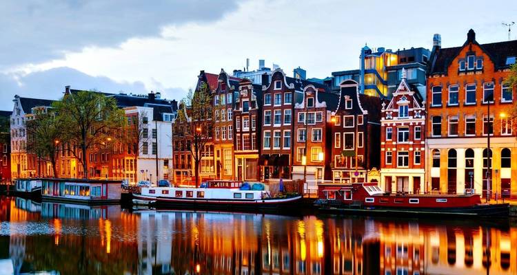 Nighttime panorama of Amsterdam’s gabled houses reflecting on tranquil canal waters with warm lights.