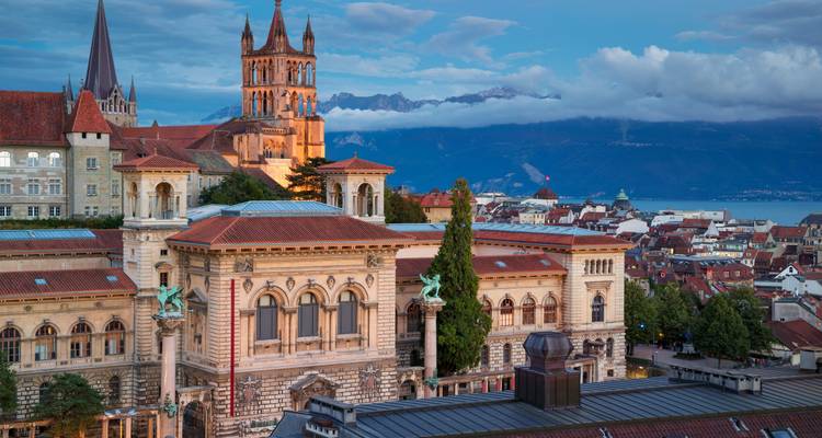 Vue du soir de la Cathédrale de Lausanne illuminée au-dessus des toits avec les Alpes et le lac en arrière-plan