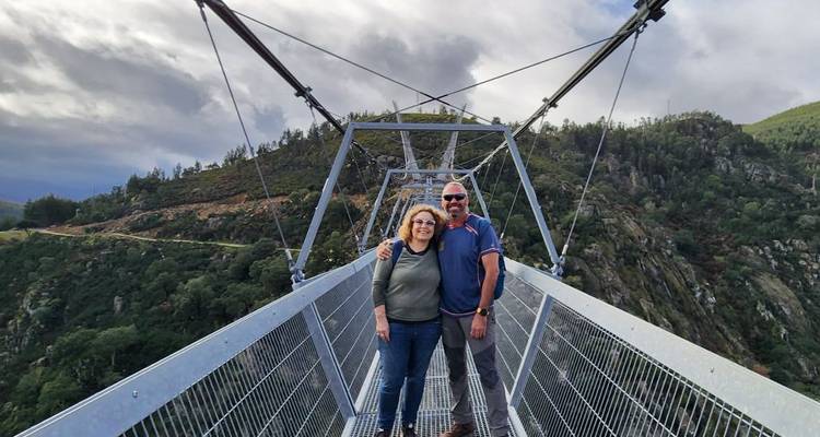 Smiling couple stands on a dramatic steel suspension footbridge high above a forested gorge.