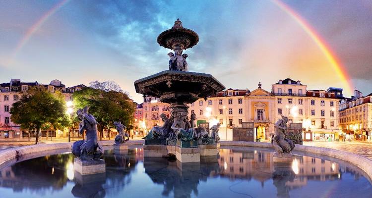 Rossio Square fountain reflected in still water beneath a glowing rainbow at twilight in Lisbon.