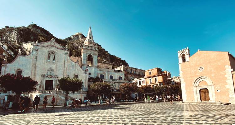 Sunny plaza in Taormina with historic churches, mountains behind and chequered stone square