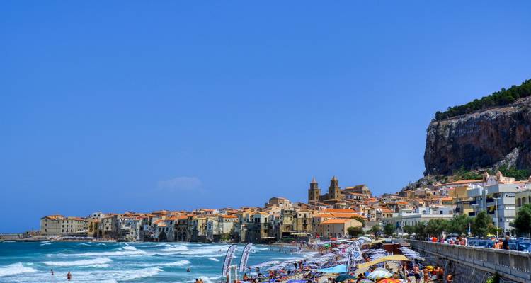 Busy beach scene at Cefalù with waves, umbrellas and medieval town along shoreline