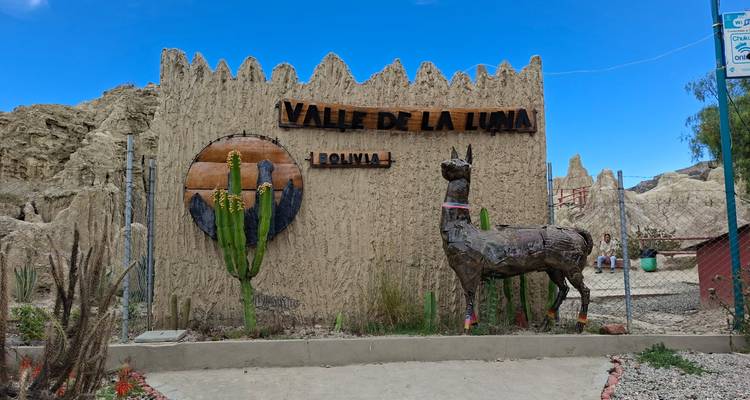 Entrance sign for Valle de la Luna in Bolivia made of adobe with cacti and a metal llama sculpture against a blue sky.