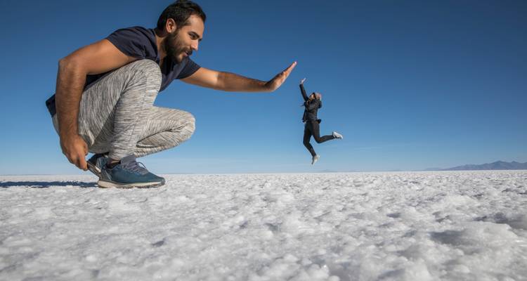 Playful perspective shot on the white expanse of the Uyuni salt flats with one person crouching and another appearing tiny in mid-air high-five pose.