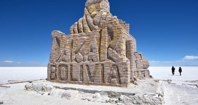 Huge salt monument reading 'DAKAR BOLIVIA' rising from the blindingly white Salar de Uyuni with two tourists walking nearby.
