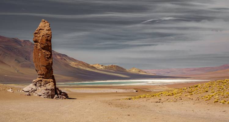 Een eenzame torenhoge stenen zuil rijst op uit een uitgestrekte hooggelegen woestijn met veelkleurige heuvels en een turquoise lagune in de verte.