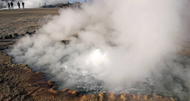 Dikke stoom walmt op uit een borrelende geothermische poel terwijl bezoekers in de verte het geysserveld observeren.