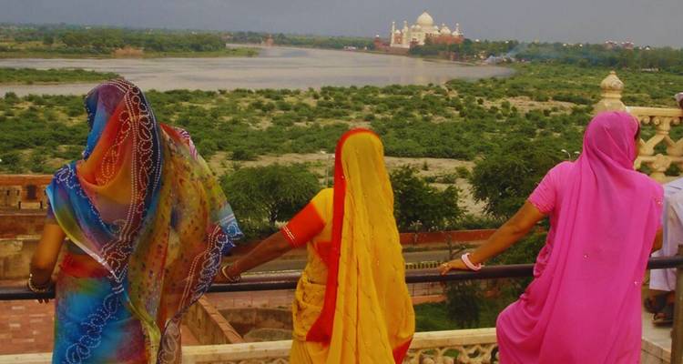 Three women in bright saris overlook the Yamuna River with the distant Taj Mahal framed in haze.