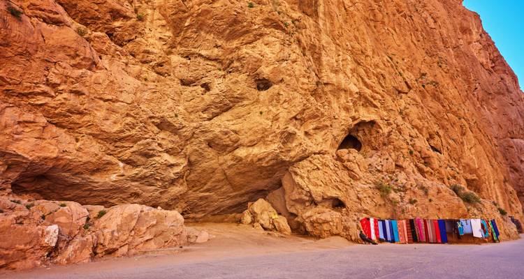 Fuß einer hoch aufragenden orangefarbenen Klippe mit bunten Teppichen zum Verkauf am Straßenrand in der Todra-Schlucht.
