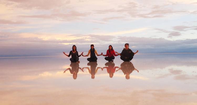 Vier vrienden zitten kleermakerszit met perfecte reflecties op de spiegelgladde Uyuni zoutvlakte tijdens pastelkleurige zonsondergang.