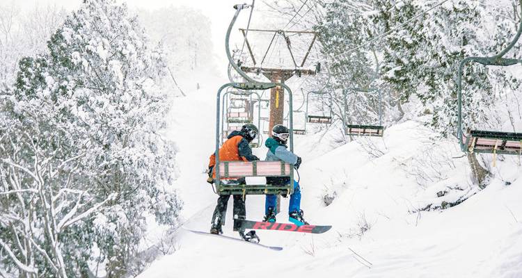 Skifahrer fahren mit einem Sessellift über schneebedeckte Bäume auf einem pulvrigen Winterhang.