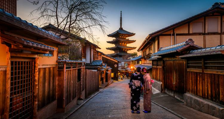 Zwei Frauen in Kimonos wandeln durch eine laternenbeleuchtete historische Gasse in Richtung Yasaka-Pagode bei Abenddämmerung in Kyoto.