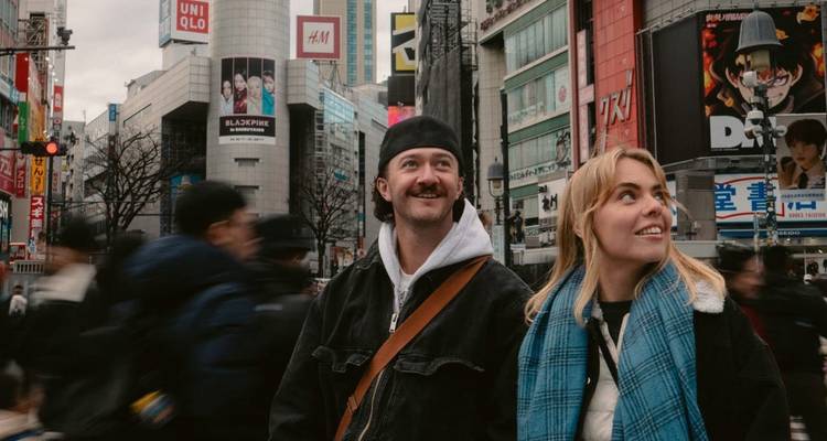 Couple smiles amid bustling crowds at Tokyo's Shibuya Crossing with neon signs above.