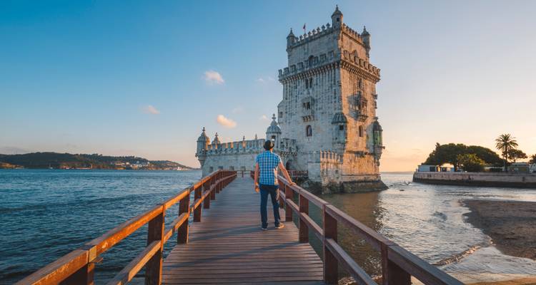 Un hombre camina por un muelle de madera hacia la histórica Torre de Belém que se alza sobre el río Tajo al atardecer.