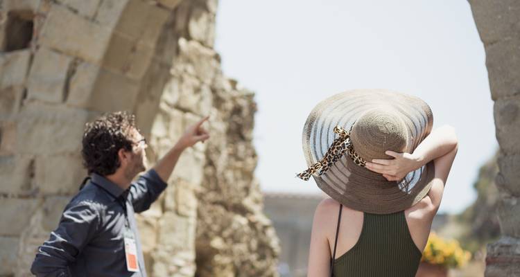 Un guide pointe vers d'anciennes ruines de pierre tandis qu'une femme avec un chapeau mou observe, encadrée par des arches usées par le temps.