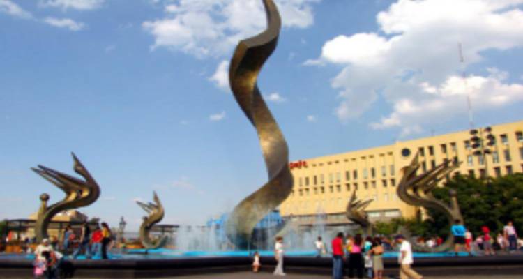 Moderne sculpturale fontein met spiraalvormige zuil en bronzen vogels omringd door bezoekers op een plein.