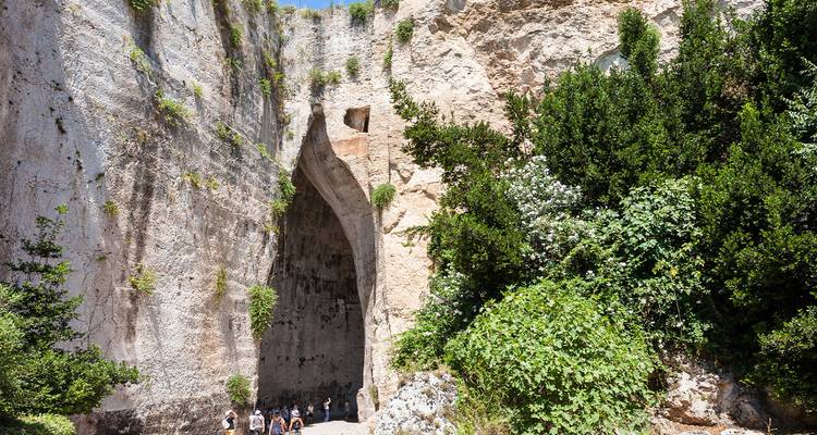 Los visitantes se encuentran en la base de la masiva cueva de piedra caliza Oreja de Dionisio en Siracusa, rodeados de vegetación.