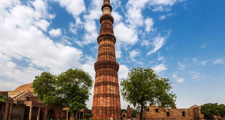 der hoch aufragende Qutub Minar, der in einen teilweise bewölkten blauen Himmel ragt, flankiert von grünen Bäumen