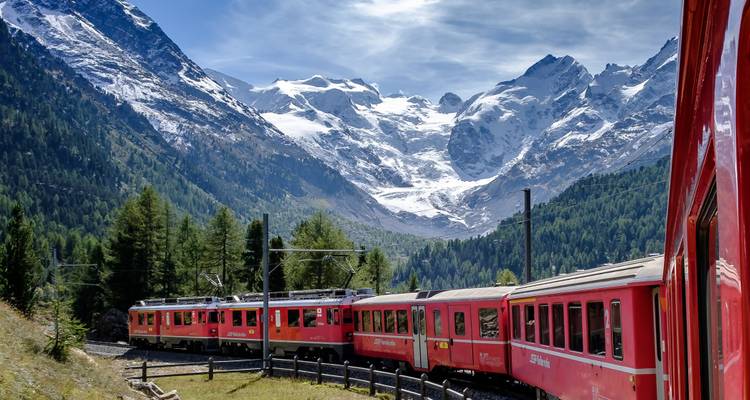 Rode panoramische trein slingert door dennenbossen met torenhoge sneeuwbedekte toppen op de achtergrond.