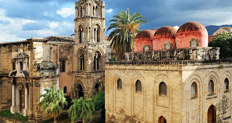 Complexe d'église historique de Palerme avec clocher orné, palmiers et dômes roses distinctifs sous un ciel dramatique.