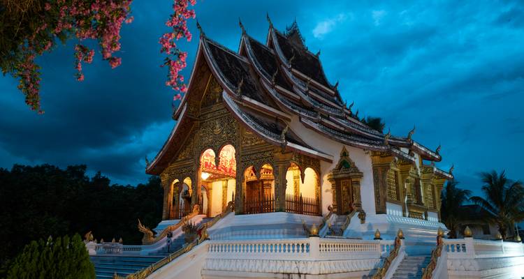 The richly decorated Wat Xieng Thong temple glows with warm interior light against a deep blue evening sky, framed by pink bougainvillea.