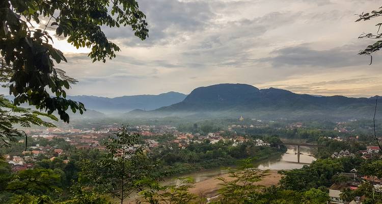 Misty dawn over Luang Prabang valley with river bends, tiled rooftops and dark silhouettes of surrounding mountains.