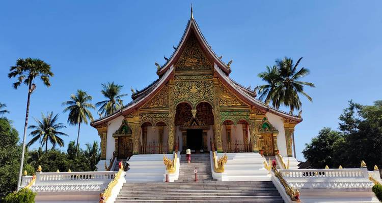 Front stairway leads to a richly ornamented Lao temple with green and gold facade surrounded by palm trees under a clear sky.