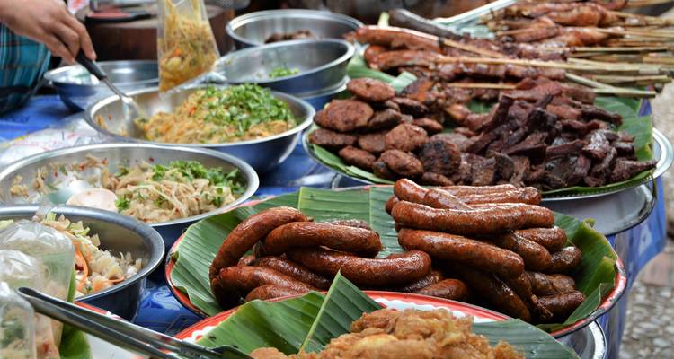 A bustling market table piled high with Lao street food including grilled sausages, meats and fresh salads on banana leaves.