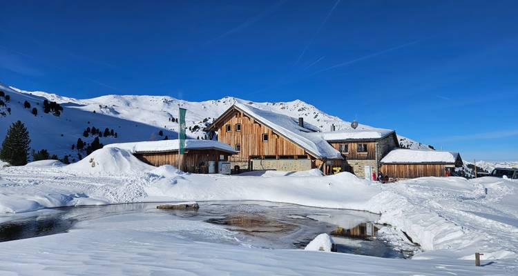Snow-blanketed mountain lodge with reflections in a small pond under bright blue skies.
