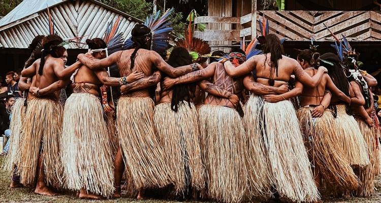 Indigenous Amazon dancers wearing straw skirts and feathered headdresses embrace in a circle.