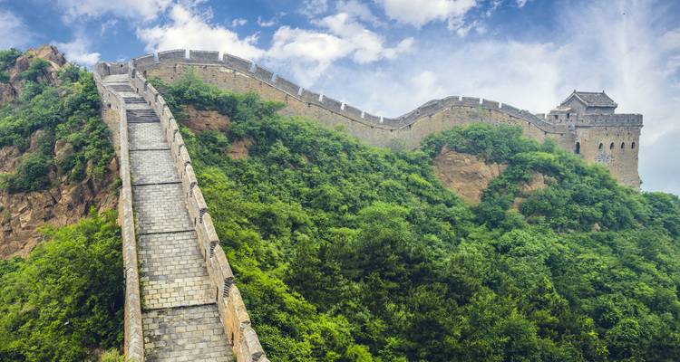 Steep stone steps climbing along a lush green section of the Great Wall of China against a bright sky.