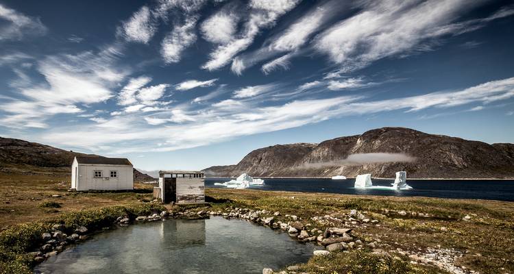 Abgelegene Küstenlandschaft mit einem kleinen weißen Häuschen und einer Holzhütte neben einem klaren Teich, mit Blick auf eine Bucht voller Eisberge, umgeben von schroffen Bergen unter einem strahlend blauen Himmel mit streifigen Wolken.