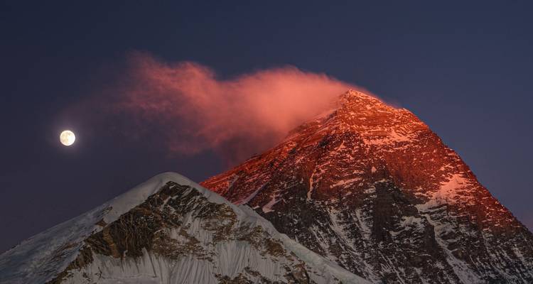 Schneebedeckter Gipfel, der im Morgengrauen rot leuchtet, mit einem Vollmond daneben vor tiefblauem Himmel