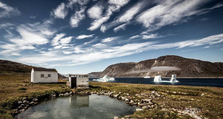 Ländliche Fjordlandschaft mit kleinen Seen, von Eisbergen gespickter Bucht und rauen Bergen unter gestreiftem Himmel.