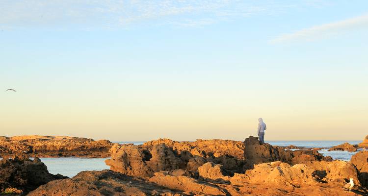 Figura solitaria se alza sobre rocas costeras escarpadas bañadas en cálida luz vespertina con mar en calma al fondo