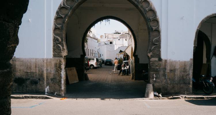 Ein steinerner Torbogen rahmt eine ruhige Gasse ein, in der geparkte Autos stehen und einige Einheimische im Schatten der weiß getünchten Medina-Mauern sitzen und sich unterhalten.