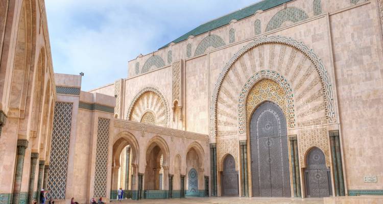 Detaillierte Fassade der Hassan-II-Moschee mit ihren riesigen, kunstvoll verzierten Türen und einem muschelförmigen Bogen, einige wenige Besucher, die von der Architektur in den Schatten gestellt werden.