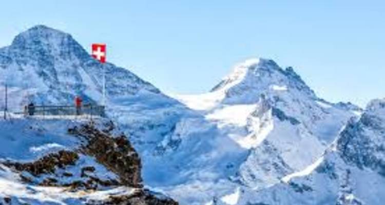 Snow-covered alpine peaks with a Swiss flag fluttering near a cliff-edge viewpoint.