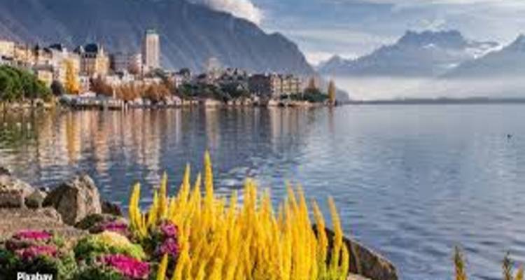 Flower-lined promenade beside a calm lake with Montreux town and distant snowy peaks.