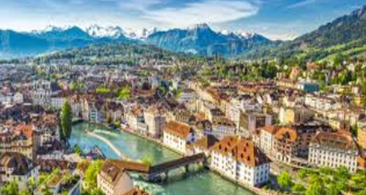 Colorful rooftops and river spanned by a covered bridge with snow-capped peaks behind.