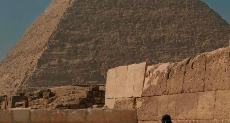 Besucher geht neben verwitterter Steinmauer mit der Großen Pyramide, die dahinter aufragt