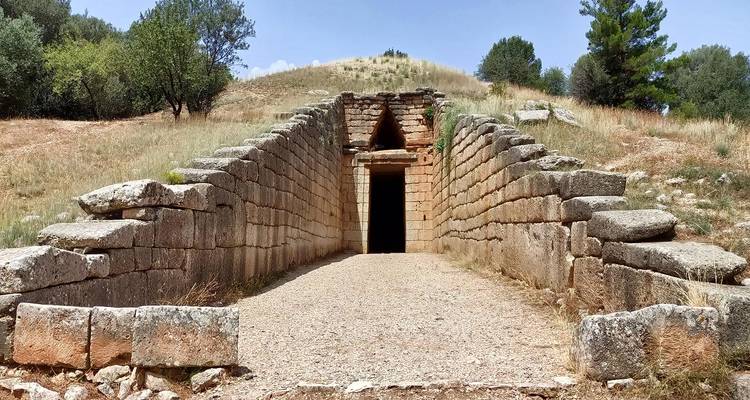 Un étroit passage de pierre mène à l'entrée sombre d'une tombe tholos mycénienne située dans des prairies sèches.