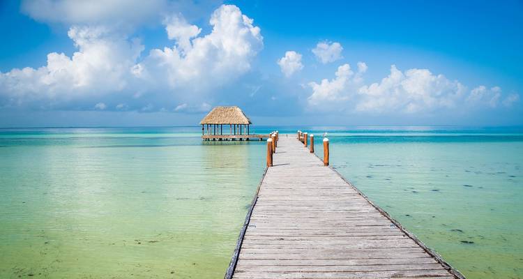 Ein langer Holzsteg, der zu einer strohgedeckten Hütte über kristallklarem türkisfarbenem Wasser unter einem strahlend blauen Himmel mit fluffigen Wolken führt.