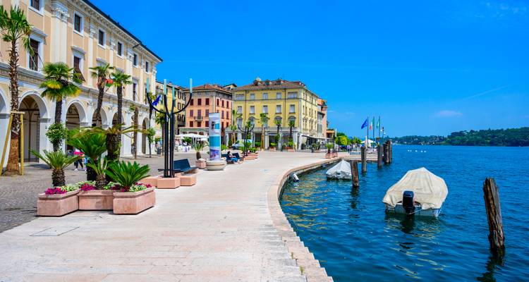 Promenade ensoleillée au bord de l'eau avec des bâtiments pastel et des bateaux amarrés le long du lac de Garde bleu.