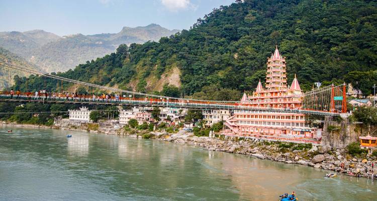 Ein leuchtend rosafarbener Tera Manzil Tempel und eine Hängebrücke überspannen den Ganges unterhalb bewaldeter Hügel in Rishikesh.