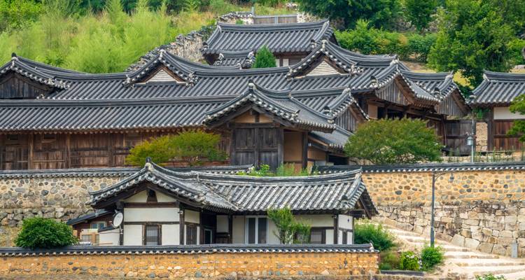 Groupe de maisons traditionnelles aux toits de tuiles dans un village entouré de murs de pierre sur fond de verdure luxuriante.