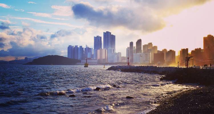 Horizon moderne de Busan s'élevant au-dessus d'un rivage rocheux avec des vagues dans la lumière dorée du soir.